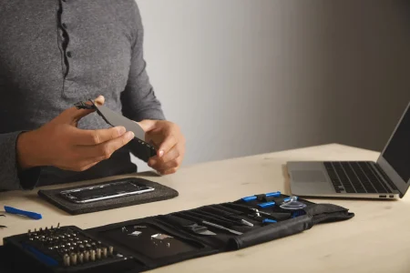 Computer and phone repairment service Master holds new screen for replacement above disassembled smartphone in his laboratory, tool kit with instruments and laptop in front of him on white table, space for your text on right